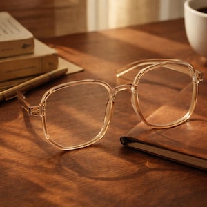 May include: Clear-framed eyeglasses with square lenses rest on a brown leather-bound notebook. A pen, books, and a cup of coffee are also on the wooden surface. The books have the titles "Joan Ingles" and "LOVE THEREAFS".