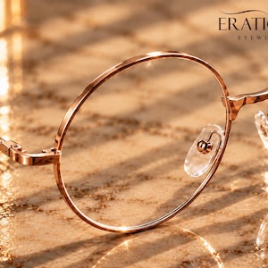 May include: Rose gold metal eyeglasses with thin, round frames and clear nose pads. The brand name ERATIQUE EYEWEAR is printed in the upper right corner. The glasses feature a metallic finish that reflects light, highlighting the circular rims.