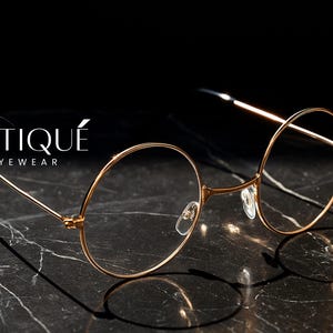 May include: Gold-rimmed eyeglasses with a round frame, resting on a dark marble surface. The glasses have a delicate, minimalist design, with the brand name "ERATIQUE EYEWEAR" in white script. The image is set against a black background.