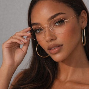 May include: A woman wearing gold-rimmed, oval eyeglasses. The glasses are held up by the hand. The image also shows gold hoop earrings and the text "ERATIQUE EYEWEAR".