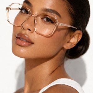 May include: A close-up of a person wearing clear-framed eyeglasses with a square shape. The glasses have a light brown tint and are set against a white background. The person is wearing a white tank top.