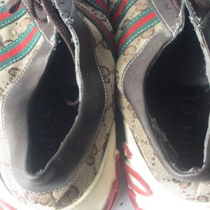Puede incluir: Par de zapatillas Gucci. Los zapatos presentan un patr&oacute;n de monograma marr&oacute;n y beige con rayas rojas y verdes. El interior del zapato es marr&oacute;n y la suela es blanca con detalles en rojo. La palabra "Gucci" es visible dentro del zapato.