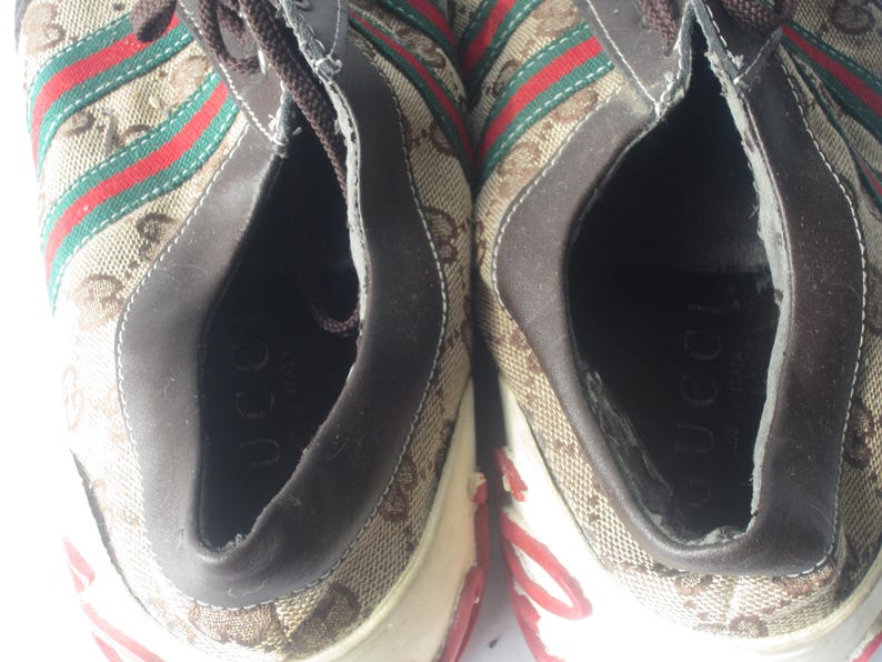 Puede incluir: Par de zapatillas Gucci. Los zapatos presentan un patr&oacute;n de monograma marr&oacute;n y beige con rayas rojas y verdes. El interior del zapato es marr&oacute;n y la suela es blanca con detalles en rojo. La palabra "Gucci" es visible dentro del zapato.