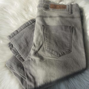 May include: A pair of light gray denim jeans, folded and resting on a white, fluffy surface. The jeans feature a classic five-pocket design, with a back pocket visible. A brown leather patch is visible on the waistband.