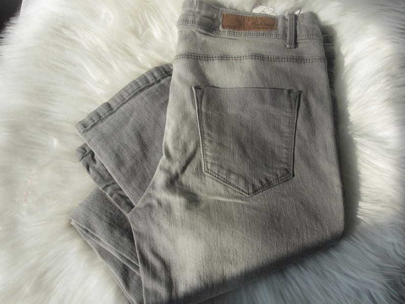 May include: A pair of light gray denim jeans, folded and resting on a white, fluffy surface. The jeans feature a classic five-pocket design, with a back pocket visible. A brown leather patch is visible on the waistband.