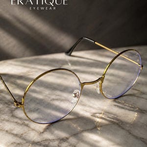May include: Gold-rimmed eyeglasses with round lenses. The glasses have a thin gold frame and black temples. The brand name "ERATIQUE EYEWEAR" is displayed in white text at the top of the image. The glasses are resting on a marble surface.
