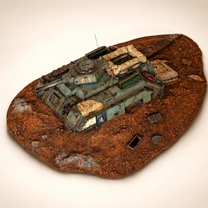 May include: A detailed model tank, painted in blue and green, rests on a textured, brown terrain base. The tank has a long gun barrel, sandbag structures, and the number "611". The base has a rough, earthy appearance, reminiscent of a battlefield.