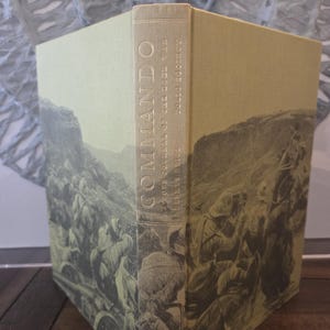 Peut inclure: Un livre à couverture rigide intitulé "Commando: A Boer Journal of the Boer War" avec une illustration sépia de soldats. Le dos comporte le titre et le nom de l'auteur en lettres dorées. Le livre a une couverture vert clair.