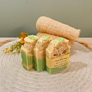 May include: Three bars of goat milk soap with a loofah sponge, and a sunflower. The soap is green and cream colored, with a loofah sponge attached. The soap has a yellow label that reads "Goat Milk Soap with tallow Natural & Organic."