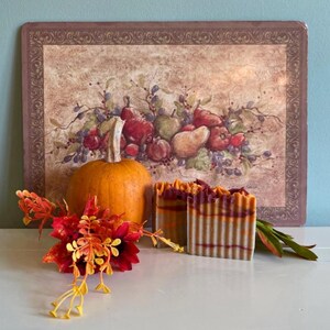 May include: Two bars of handmade soap with orange, brown, and white swirls. The soap is sitting on a table next to a small orange pumpkin and a fall-themed floral arrangement.
