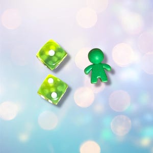 K&ouml;nnte beinhalten: Zwei gr&uuml;ne W&uuml;rfel mit wei&szlig;en Punkten und eine gr&uuml;ne Figur auf einem hellblauen Hintergrund mit wei&szlig;en und rosa Bokeh.