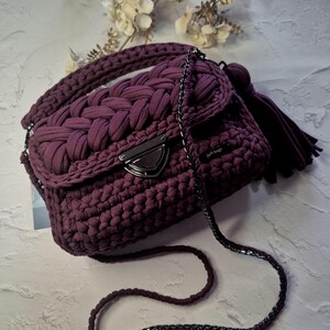 May include: A plum-colored, handmade crochet handbag with a braided top handle and a long chain strap. The bag features a flap closure with a silver-toned clasp and a matching tassel. The bag is a stylish accessory.