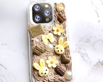 Puede incluir: Una funda transparente para teléfono con un tema de chocolate y conejito marrón y blanco. La funda está decorada con barras de chocolate, corazones de chocolate, flores blancas, perlas blancas y dos figuritas de conejito amarillas.
