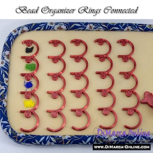 May include: Red bead organizer with 26 connected rings labeled with letters from A to Z. Each ring has a small compartment for beads. The organizer is on a blue and white floral fabric background.