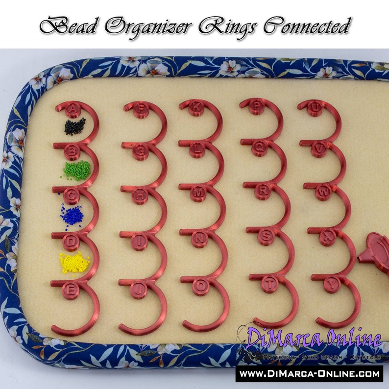 May include: Red bead organizer with 26 connected rings labeled with letters from A to Z. Each ring has a small compartment for beads. The organizer is on a blue and white floral fabric background.