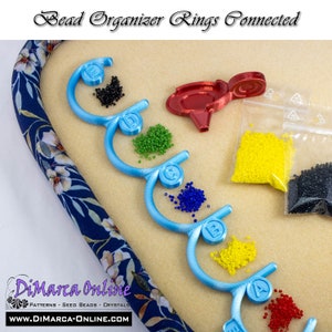 May include: Blue bead organizer rings with letters A, B, C, D, and E. Each ring holds a different color of seed beads: red, yellow, blue, green, and black. The rings are connected together and are on a beige surface.