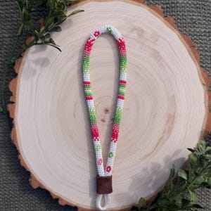 May include: A beaded wrist lanyard featuring a repeating pattern of small white, lime green, and bright pink beads. The beads are arranged in a continuous loop, secured at the base by a brown suede-like fabric connector with a white cord loop.