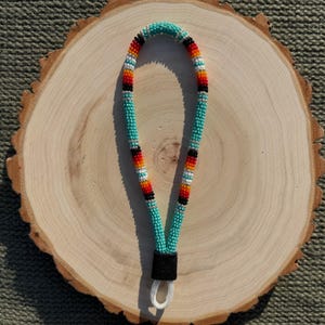 May include: A beaded lanyard with a turquoise background and colorful stripes of red, orange, white, and black. The lanyard is made of small beads and has a black leather piece at the bottom with a white loop.