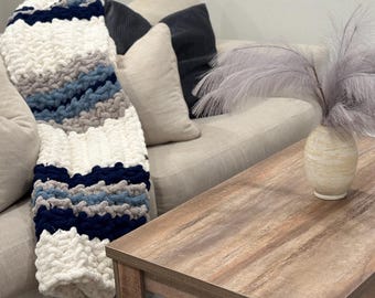 Könnte beinhalten: Ein weißes Sofa mit einer gestrickten blauen, grauen und weißen Wolldecke. Vor dem Sofa steht ein Holz-Couchtisch mit einer Vase mit Trockenblumen.