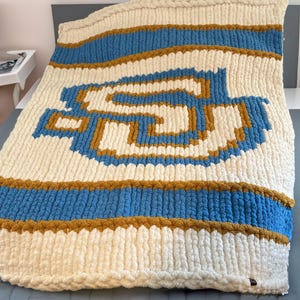 Puede incluir: Una manta de punto de color crema con un diseño geométrico azul y dorado. La manta tiene un borde trenzado y está doblada en una cama.