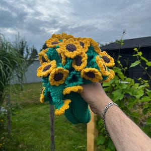 Könnte beinhalten: Ein handgefertigter Häkelstrauß Sonnenblumen. Die Sonnenblumen haben gelbe Blütenblätter mit braunen Zentren und sind von grünen Blättern umgeben. Der Strauß wird in einer grünen gehäkelten Basis gehalten.