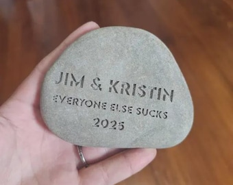 May include: A smooth, light gray river stone with dark engraved text. The inscription reads JIM & KRISTIN, followed by the phrase EVERYONE ELSE SUCKS, and the year 2025 at the bottom. The stone has a rounded, irregular shape and a matte texture.