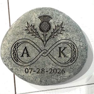 May include: A smooth, gray, rounded river stone featuring a laser-engraved design. The engraving includes a thistle flower above an infinity symbol containing the letters A and K, with the date 07-28-2026 inscribed at the bottom.
