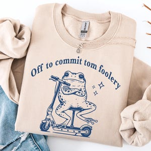 Könnte beinhalten: Beiges Sweatshirt mit dem marineblauen Schriftzug "Off to commit tom foolery". Die Grafik zeigt einen Frosch auf einem Roller. Das Sweatshirt ist neben einer blauen Jeans gefaltet.