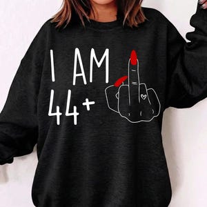 Op de afbeelding: Zwart sweatshirt met de witte tekst "I AM 44+". Aan de rechterkant een witte omtrek van een hand met een opgestoken middelvinger en rode nagellak. Op de hand is een klein hartje getekend.
