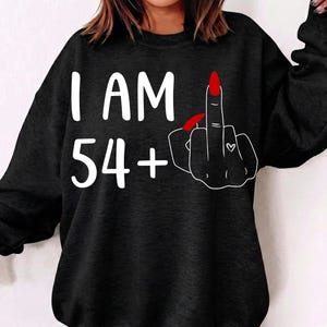 Könnte beinhalten: Schwarzes Sweatshirt mit dem weißen Text "I AM 54+". Eine Hand mit rotem Fingernagel zeigt den Mittelfinger, mit einem kleinen Herz auf dem Ringfinger.