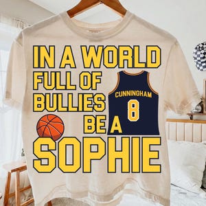 Op de afbeelding: Een crèmekleurig T-shirt met korte mouwen met de gele tekst IN A WORLD FULL OF BULLIES BE A SOPHIE, een afbeelding van een oranje basketbal en een marineblauw basketbalshirt met de naam CUNNINGHAM en het nummer 8 in het geel.