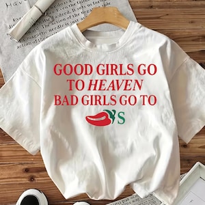 May include: White t-shirt featuring the text "GOOD GIRLS GO TO HEAVEN BAD GIRLS GO TO" in red, and a red chili pepper graphic with green leaves. The shirt is displayed on a wooden hanger.