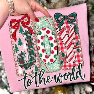 May include: A light pink t-shirt featuring a graphic with the word JOY in large letters, each with a different pattern: green plaid, red and green polka dots, and red and white stripes. The text to the world is printed below in dark green.