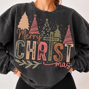 Puede incluir: Una sudadera gris carbón con efecto lavado al ácido que presenta un estampado gráfico con el texto Merry CHRISTmas, donde la letra I se sustituye por una cruz. El diseño incluye pinos estilizados en tonos rosa, beige y verde oliva.