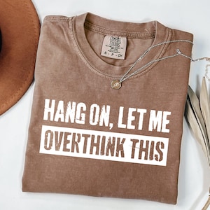 Op de afbeelding: Een bruin Comfort Colors t-shirt met de tekst "HANG ON, LET ME OVERTHINK THIS" in witte blokletters. Een zilveren ketting met een kleine hanger ligt over het shirt. Een bruine hoed is zichtbaar in de linkerbovenhoek.