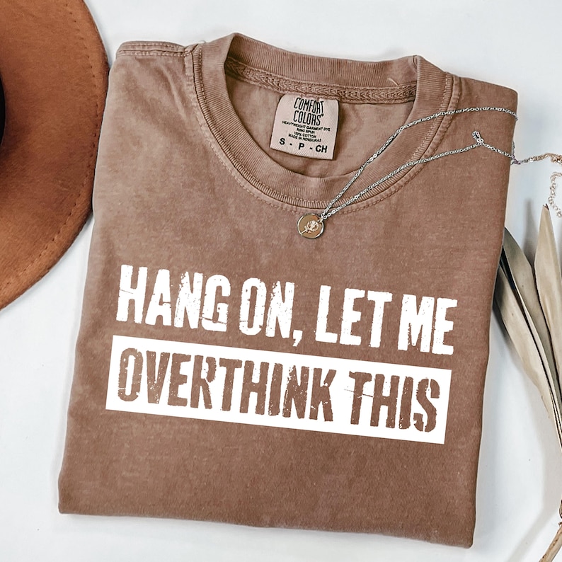 Op de afbeelding: Een bruin Comfort Colors t-shirt met de tekst "HANG ON, LET ME OVERTHINK THIS" in witte blokletters. Een zilveren ketting met een kleine hanger ligt over het shirt. Een bruine hoed is zichtbaar in de linkerbovenhoek.