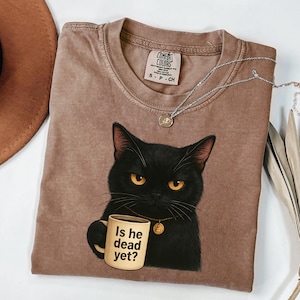 May include: A brown Comfort Colors t-shirt featuring a black cat with yellow eyes holding a mug. The mug reads "Is he dead yet?" A silver necklace with a pendant is draped on the shirt. The shirt is size S-P-CH.