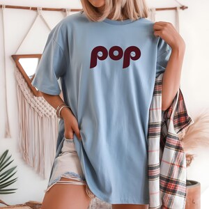 May include: A light blue t-shirt with the word "pop" in a burgundy color. The shirt is a relaxed fit and has short sleeves. A plaid shirt is draped over the shoulder.