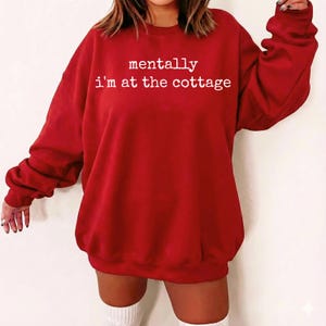 Puede incluir: Una sudadera roja con el texto "mentally i'm at the cottage" en blanco. La parte superior de manga larga es de color liso y parece estar hecha de un material suave. La sudadera es de estilo casual.