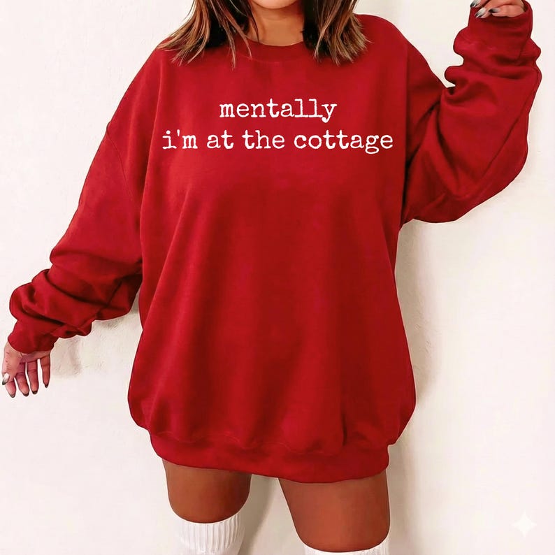 Puede incluir: Una sudadera roja con el texto "mentally i'm at the cottage" en blanco. La parte superior de manga larga es de color liso y parece estar hecha de un material suave. La sudadera es de estilo casual.