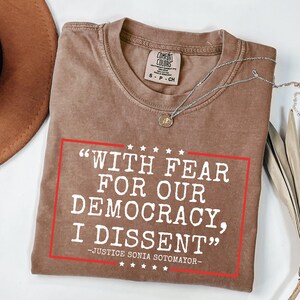 Può includere: Una maglietta marrone con il testo "WITH FEAR FOR OUR DEMOCRACY, I DISSENT" in lettere bianche. Il testo è incorniciato da un rettangolo rosso con cinque stelle bianche sopra e sotto il testo. Il testo "JUSTICE SONIA SOTOMAYOR" è sotto il rettangolo in lettere bianche più piccole.