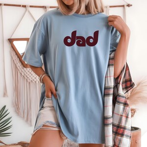 May include: A light blue t-shirt with the word "Dad" in a maroon, retro font. The shirt is a relaxed fit and is being worn with denim shorts. A plaid shirt is draped over the arm.