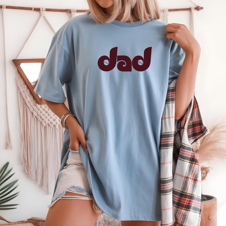 May include: A light blue t-shirt with the word "Dad" in a maroon, retro font. The shirt is a relaxed fit and is being worn with denim shorts. A plaid shirt is draped over the arm.