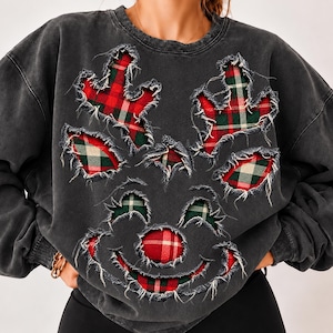 Puede incluir: Una sudadera de cuello redondo gris carbón con un diseño de aplique deshilachado en forma de cara de reno. Las astas, los ojos y la nariz del reno están rellenos con una tela de cuadros rojos, verdes y blancos sobre la tela oscura.