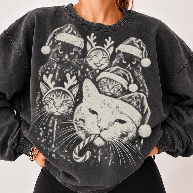 Puede incluir: Una sudadera de cuello redondo en color gris carb&oacute;n con un gran estampado gr&aacute;fico de varios gatos con gorros de Pap&aacute; Noel y astas de reno. Un gato en primer plano sostiene un bast&oacute;n de caramelo en la boca, todo en tinta blanca desgastada.