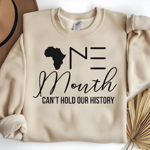 Puede incluir: Sudadera beige con el texto negro "ONE Month CAN'T HOLD OUR HISTORY". La palabra "ONE" está estilizada con la letra "N" y tres líneas horizontales. Una silueta negra del continente africano está encima de la "N".