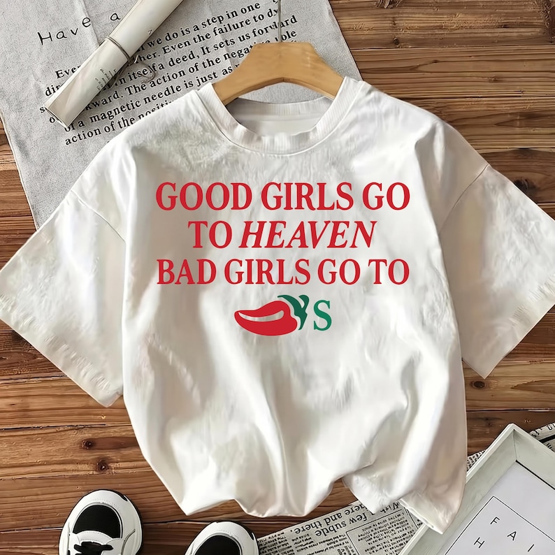 May include: White t-shirt featuring the text "GOOD GIRLS GO TO HEAVEN BAD GIRLS GO TO" in red, and a red chili pepper graphic with green leaves. The shirt is displayed on a wooden hanger.