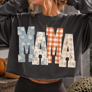 May include: A charcoal gray long-sleeve sweatshirt with a raw-edge hem. The word MAMA is printed across the front in large block letters. Each letter features a different pattern: blue leaves, blue floral, orange gingham, and plaid with pumpkins.