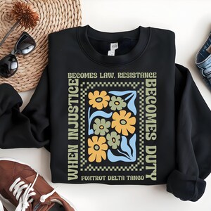 Pode incluir: Camisola de moletom preta com uma estampa gráfica de flores e o texto "When injustice becomes law, resistance becomes duty. Foxtrot Delta Tango."