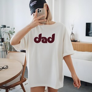 May include: Cream-colored t-shirt with the word "Dad" in a burgundy, retro-style font. The shirt has short sleeves and a relaxed fit. The person in the image is wearing a black baseball cap and holding a phone.